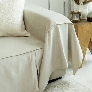 May include: A light beige sofa with a matching throw blanket draped over the back and arms. The sofa has a pleated skirt and is sitting on a light beige rug.