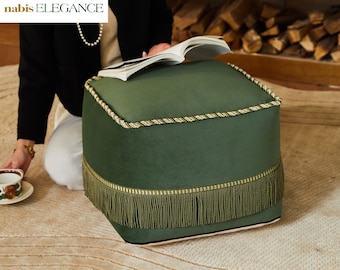 May include: A square green velvet ottoman with a gold and white rope trim and a green fringe around the bottom.