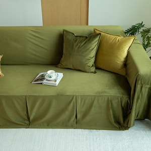 May include: Olive green velvet sofa with matching throw pillows in shades of green and gold. A book and teacup rest on the sofa. The sofa has a skirted base and is in a well-lit room.