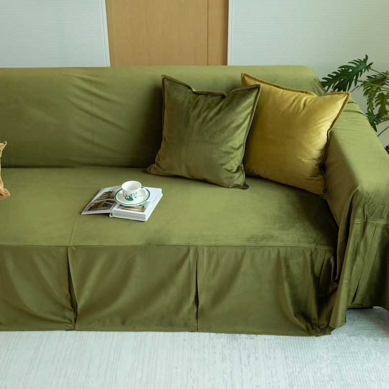 May include: Olive green velvet sofa with matching throw pillows in shades of green and gold. A book and teacup rest on the sofa. The sofa has a skirted base and is in a well-lit room.