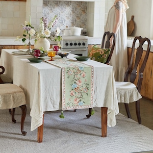 Pode incluir: Uma toalha de mesa de linho branco com um corredor de mesa com padrão floral. O corredor de mesa tem um fundo verde com flores rosa, azuis e brancas. A mesa está posta com dois talheres e um vaso de flores.