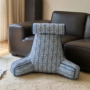 May include: A white and blue patterned wedge pillow with a backrest. The pillow has a geometric design with blue lines on a white background. The pillow is sitting on a light beige rug.