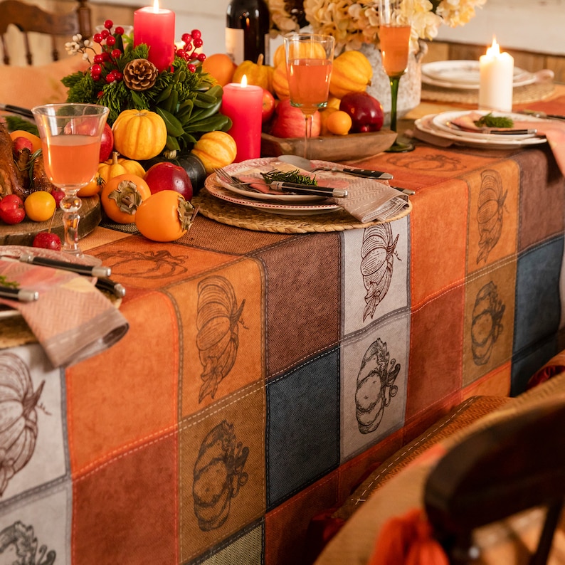 May include: Rectangular fall tablecloth with orange, brown, white, and dark green patchwork blocks, pumpkin illustrations. The table is set with candles, fruit, drinks, and plates.