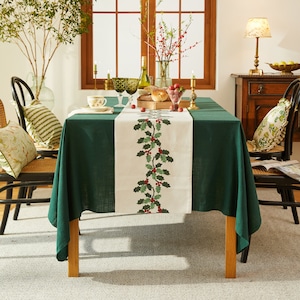 May include: A green tablecloth with a white runner featuring a holly design. The runner has a green holly leaf pattern with red berries. The tablecloth is set for a holiday dinner with two chairs and a centerpiece.
