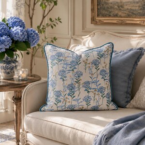 May include: A decorative square pillow with a floral hydrangea pattern in shades of blue and yellow on an off-white background. The pillow has a blue trim and is placed on a cream-colored sofa. A second blue pillow is behind it.