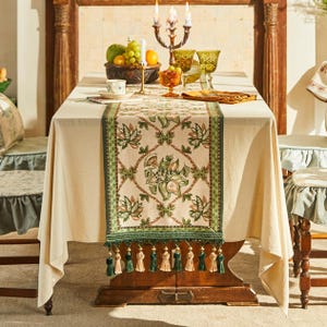 May include: A cream tablecloth with a green and cream floral table runner. The runner has a green border and tassels. A wooden table is set with fruit, candles, and glassware. The chairs have blue seat covers.