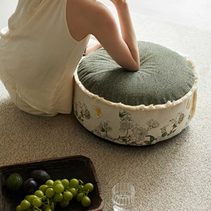 May include: A round green and white floral patterned cushion with a fringed edge. The cushion is sitting on a light beige carpet.