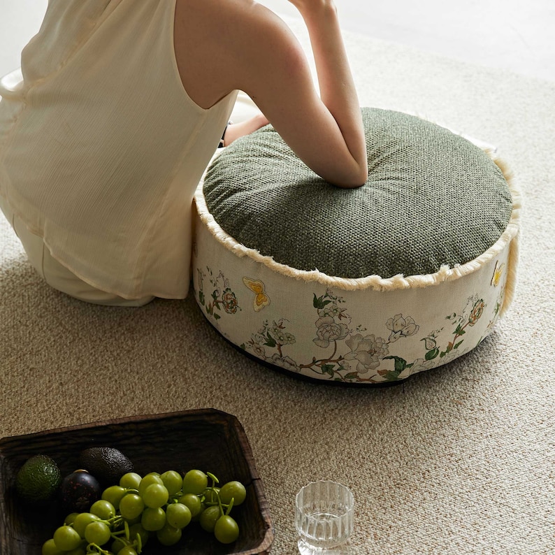 May include: A round green and white floral patterned cushion with a fringed edge. The cushion is sitting on a light beige carpet.