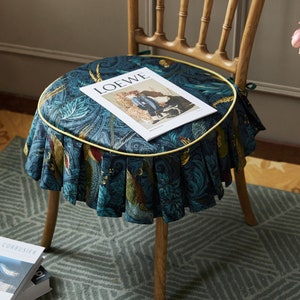 May include: A round chair cushion with a blue and green floral pattern and a yellow trim. The cushion is sitting on a wooden chair with a magazine on top that says "LOEWE".