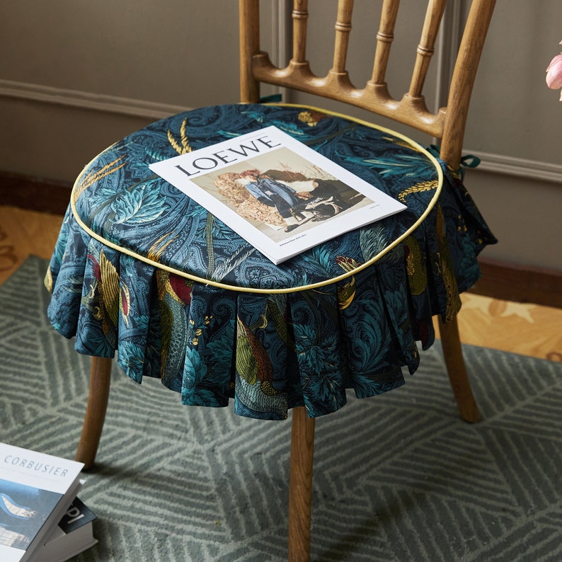 May include: A round chair cushion with a blue and green floral pattern and a yellow trim. The cushion is sitting on a wooden chair with a magazine on top that says "LOEWE".