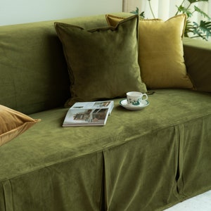 May include: A green velvet sofa with several pillows in shades of green and gold. A magazine and a teacup with a floral design sit on the sofa. The sofa has a pleated skirt.