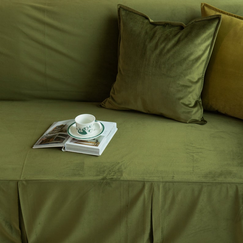 May include: A green velvet sofa with two pillows and a cup of tea on a book. The pillows are in shades of green. The cup and saucer have a green design. The sofa has a skirted base.