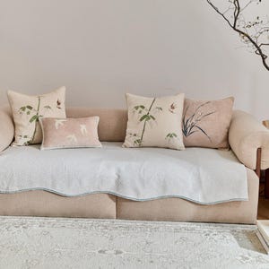 Könnte beinhalten: Ein beiges Sofa mit vier dekorativen Kissen. Zwei quadratische Kissen zeigen Bambus- und Vogelmotive, ein rechteckiges Kissen hat fliegende Vogelsilhouetten und ein weiteres ein Blumenmuster. Eine hellblaue Decke bedeckt das Sofa.