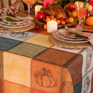 May include: A fall-themed table setting with a colorful patchwork-like tablecloth featuring pumpkin illustrations. The table is set with plates, silverware, and glasses, with a roasted chicken and fruit centerpiece.
