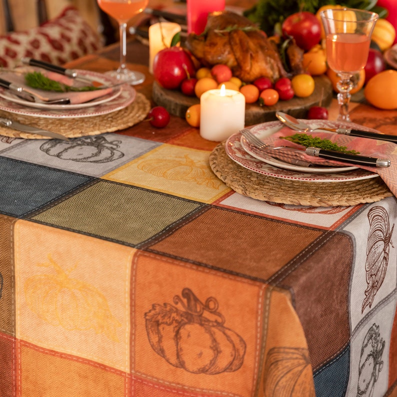 May include: A fall-themed table setting with a colorful patchwork-like tablecloth featuring pumpkin illustrations. The table is set with plates, silverware, and glasses, with a roasted chicken and fruit centerpiece.