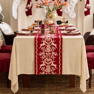 May include: A white tablecloth with a red and cream patterned table runner. The table runner features a repeating design of birds, hearts, and pine cones. The table is set for a festive dinner with glasses, plates, and cutlery.