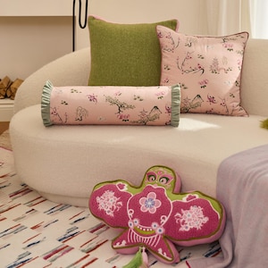 Puede incluir: Un sofá blanco con tres almohadas. Las almohadas son rosas y verdes con patrones florales. Una almohada de mariposa bordada en rosa y verde está en primer plano.