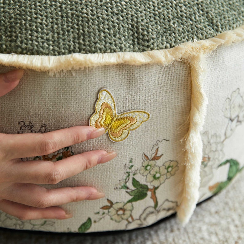 May include: A round, green and cream colored cushion with a fringe border. The cushion has an embroidered butterfly and floral design.