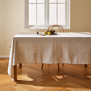 May include: A white tablecloth with a subtle floral pattern, laid on a wooden table with a few green apples and a glass on top. The table is in a room with a window and wooden floors.