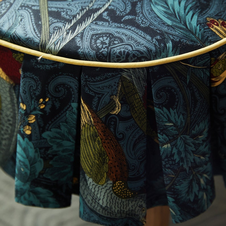 May include: A round, blue and green fabric ottoman with a yellow trim and a paisley pattern. The fabric is pleated around the bottom of the ottoman.