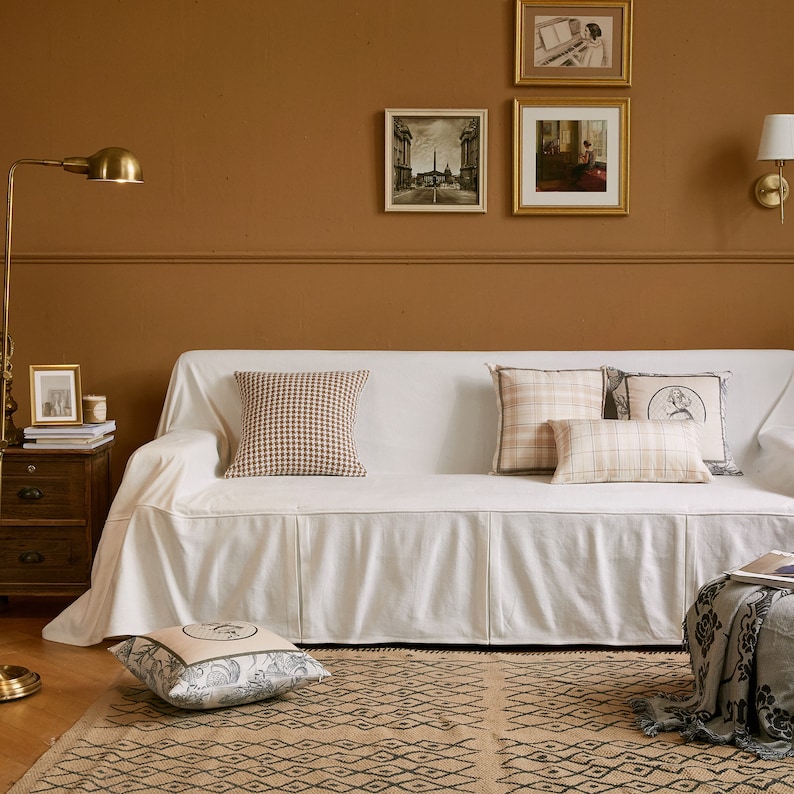 May include: A white couch with a white throw blanket and several patterned throw pillows. The couch is in a room with brown walls and a rug with a diamond pattern.
