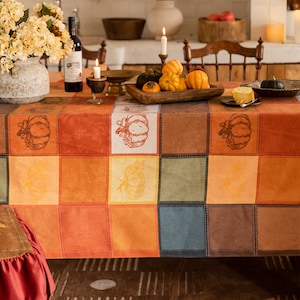 May include: A patchwork like tablecloth with a fall harvest theme. The tablecloth features a grid of squares in shades of orange, brown, and beige, with pumpkin illustrations. The table is set with candles, gourds, and a floral arrangement, creating a warm, inviting atmosphere.