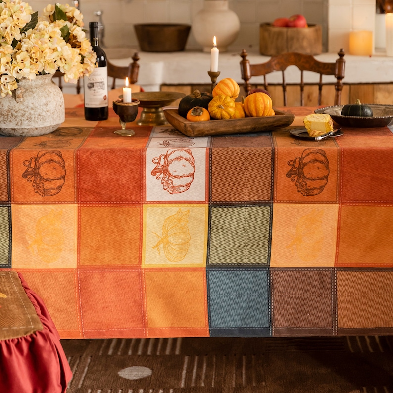 May include: A patchwork like tablecloth with a fall harvest theme. The tablecloth features a grid of squares in shades of orange, brown, and beige, with pumpkin illustrations. The table is set with candles, gourds, and a floral arrangement, creating a warm, inviting atmosphere.
