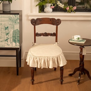 Puede incluir: Una silla de madera con respaldo tallado y una funda de silla con volantes de color crema. La silla está sentada en un suelo de madera en una habitación con una mesa auxiliar y un armario.