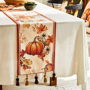 Op de afbeelding: Een beige tafelkleed met een herfstthema met pompoenen, bladeren en bloemen. De tafelkleed heeft een bruine rand en gouden kwastjes.
