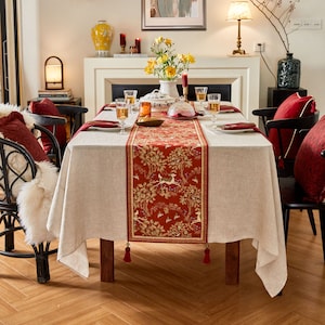 May include: A white linen tablecloth with a red and gold tapestry table runner featuring a deer design. The table is set with glasses, plates, and napkins.