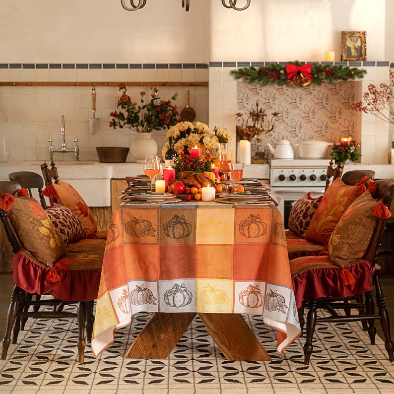 May include: Rectangular fall tablecloth with warm orange, yellow, brown, gray, and cream patchwork blocks, featuring pumpkin illustrations. The dining table is styled with candles, drinks, plates, fruit, and autumn decor.