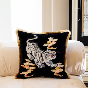 May include: A black velvet throw pillow with a white embroidered tiger and gold embroidered clouds. The pillow has a brown fringe border.