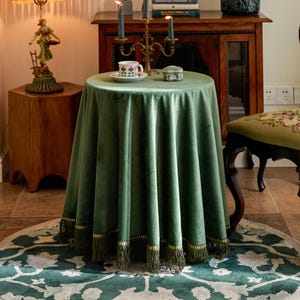 May include: A round, forest green velvet tablecloth with a fringe trim. The tablecloth is draped over a small table, with a teacup and saucer, and a small decorative box on top. The table is set in a room with antique furniture.