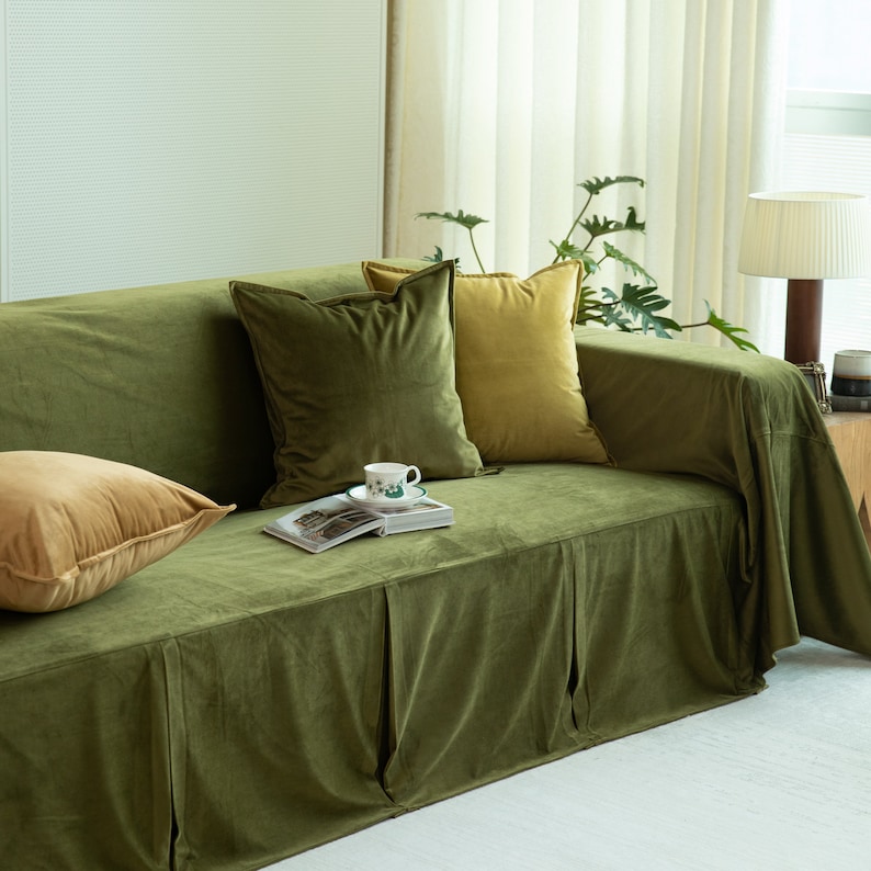 May include: A green velvet sofa with several throw pillows in shades of green and gold. A book and teacup rest on the sofa. The sofa has a skirted base and is in a well-lit room.