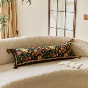 Puede incluir: Una almohada larga y rectangular con un patrón floral en tonos verdes, rojos y amarillos sobre un fondo negro. La almohada tiene borlas en cada extremo y está sentada en un sofá blanco.