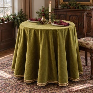 May include: A round olive green velvet tablecloth with a decorative gold braided trim along the hem. The fabric drapes to the floor, covering a circular table set with white plates, burgundy napkins, and a brass candle holder.