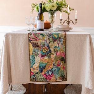 Pode incluir: Um caminho de mesa retangular com um padrão de tapeçaria multicolorido e vibrante de várias aves tropicais, incluindo papagaios e tucanos, com detalhes tecidos intrincados em tons de azul-petróleo, rosa, amarelo e verde, com acabamento em borda escura.