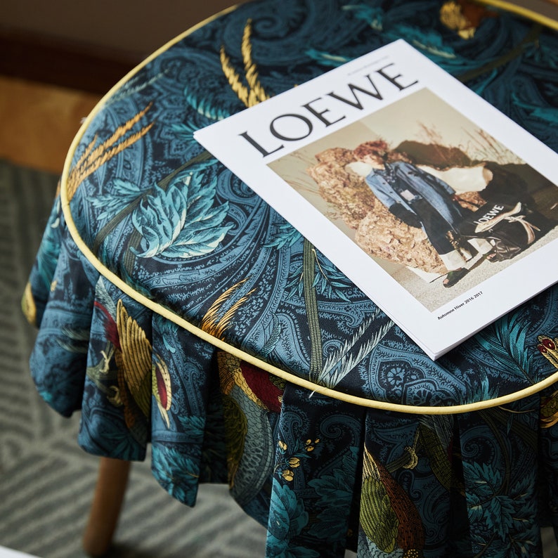 May include: A blue and gold patterned fabric stool with a yellow trim. A magazine with the word "LOEWE" printed on the cover is resting on the stool.