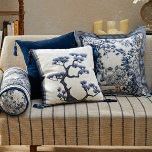 Op de afbeelding: Drie decoratieve kussens met blauwe en witte patronen. De kussens liggen op een gestreepte bank met een bruin-wit kleurenschema. De kussens tonen een dennenboom, een blauw en wit landschap en een effen blauwe kleur.