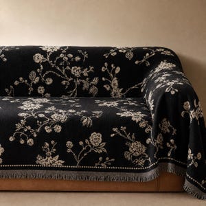 Può includere: Una coperta nera con un motivo floreale color crema caratterizzato da dettagliate illustrazioni botaniche di fiori, foglie e steli. Il tessuto presenta una trama intrecciata e un bordo decorativo con una linea tratteggiata e frange.