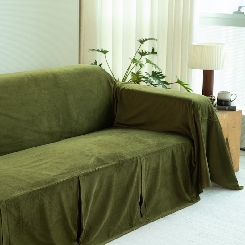 May include: Olive green velvet sofa with a skirted base. The sofa is in a well-lit room with a white rug and sheer curtains. A wooden side table holds a lamp and a mug.