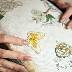 May include: A white cushion with embroidered floral designs and a yellow butterfly applique. The cushion has a fringe border.