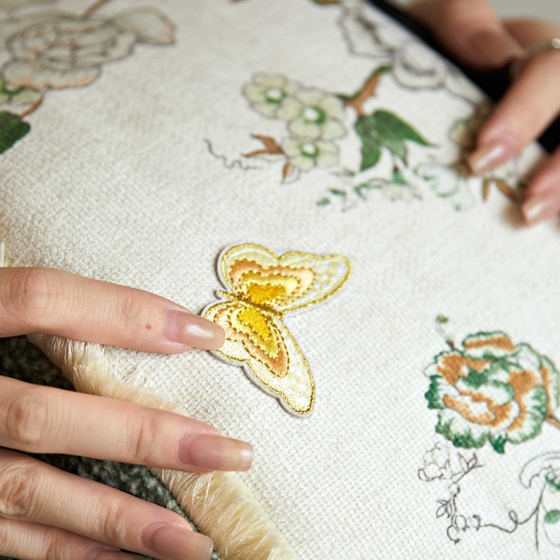 May include: A white cushion with embroidered floral designs and a yellow butterfly applique. The cushion has a fringe border.