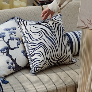May include: A white and blue striped throw pillow with a fringe border. The pillow is on a couch with a striped fabric.