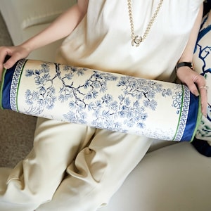 May include: A white cylindrical pillow with a blue and green floral pattern. The pillow has a border with a repeating geometric design.