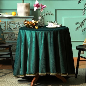 Puede incluir: Un mantel redondo de terciopelo verde esmeralda con un ribete dorado y crema. La mesa está decorada con un cuenco decorativo, una taza de té y un jarrón con flores. La mesa está sostenida por patas de madera.