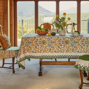 Puede incluir: Un mantel blanco con un borde a cuadros verde y negro y un patrón floral en tonos marrones, amarillos y azules. El mantel está puesto sobre una mesa de madera con un cuenco de limones y una jarra sobre la mesa.