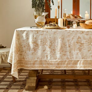 Puede incluir: Un mantel crema con un diseño de escena de bosque marrón, sobre una mesa de madera. La mesa está puesta con platos con borde dorado, una jarra de cristal, velas y una cesta de pan. El mantel tiene un borde detallado.
