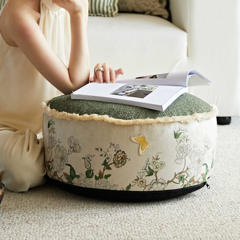 May include: A round green and cream floral patterned ottoman with a black zipper and a yellow butterfly embroidered on the front.