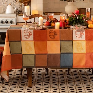 Puede incluir: Un mantel de patchwork con temática de cosecha otoñal. El patrón cuadrado presenta varios tonos de naranja, marrón y verde, con ilustraciones de calabazas. La mesa está puesta para una comida festiva, con velas, comida y bebidas.